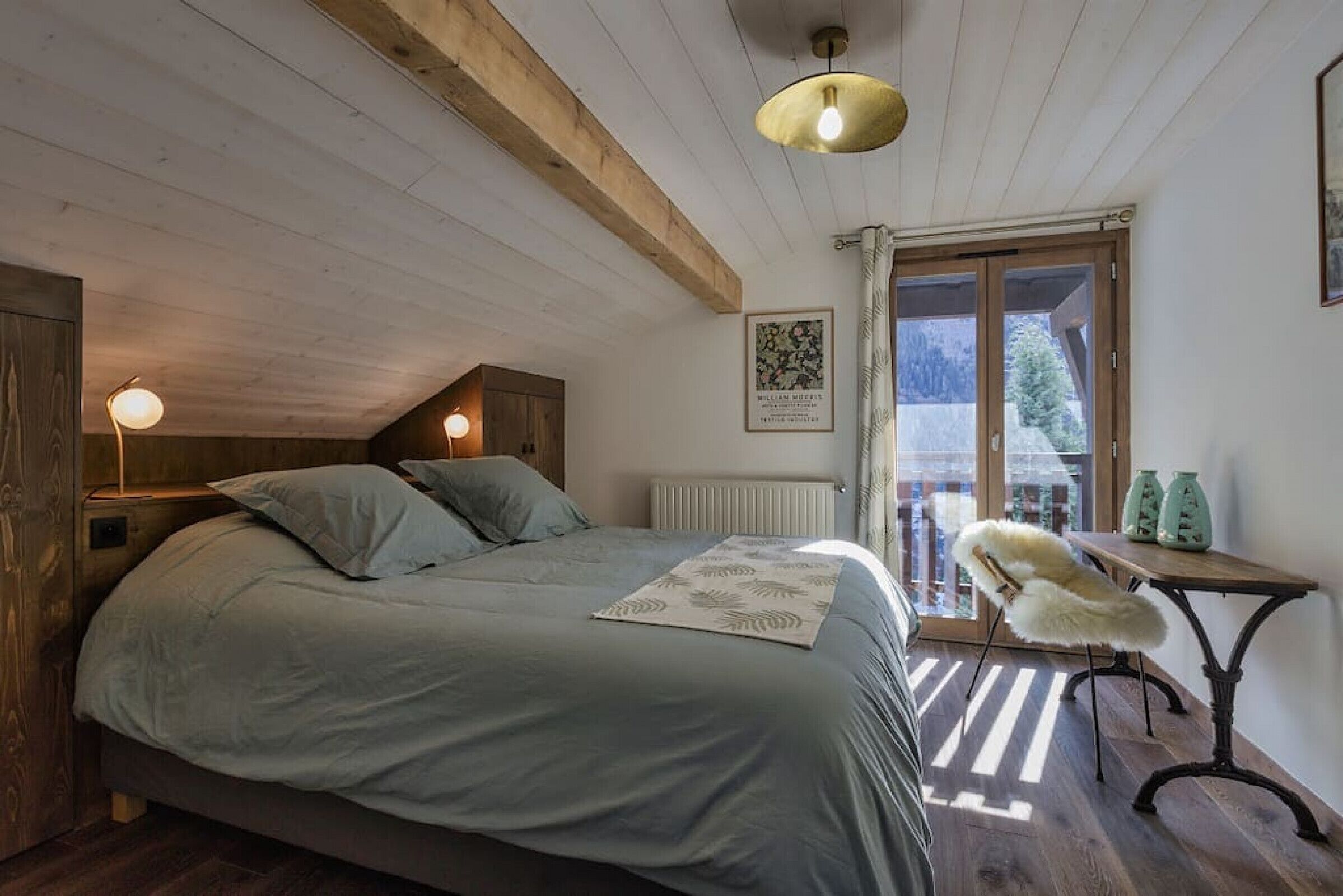A bedroom with a bed, a wooden ceiling, a radiator, a window with curtains, a small table with a chair, and a framed picture on the wall.