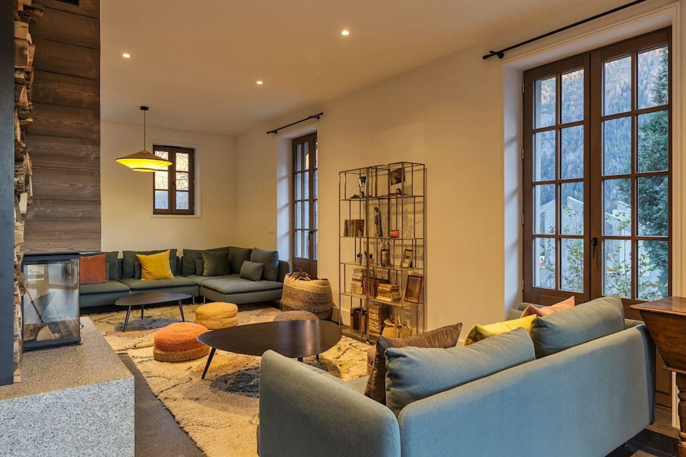 A modern living room with a sofa, ottomans, a coffee table, a bookshelf, and a large window.