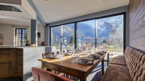 Une cuisine moderne avec une grande table à manger pouvant accueillir six personnes, un banc en bois et une vue sur les montagnes grâce à de grandes fenêtres.