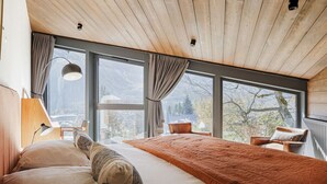 Une chambre avec un grand lit, un bureau et une chaise. Plusieurs fenêtres offrent une vue sur l’extérieur.