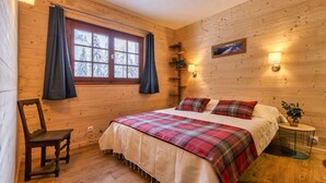 Une chambre confortable dans un chalet en bois, avec un lit, une chaise, une fenêtre avec des rideaux, une étagère fixée au mur et une petite table avec une lampe.