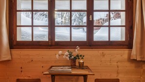 Une pièce chaleureuse au décor intérieur en bois, une fenêtre donnant sur un paysage enneigé, une petite table avec un livre et un vase de fleurs, et deux fauteuils rouges confortables.