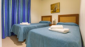 Ein Hotelzimmer mit zwei Betten, jeweils mit blauer Bettdecke und weißen Handtüchern. An der Wand hängt ein Gemälde, und am Fenster ist ein Vorhang mit blauen und grünen Streifen.