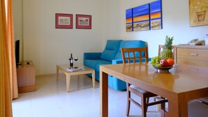 Ein Wohnzimmer mit einem blauen Sofa, einem Holzesstisch mit Stühlen, einem kleinen Couchtisch und einer Obstschale. An der Wand hängen gerahmte Bilder, ein Fernseher und ein Schreibtisch mit Telefon.