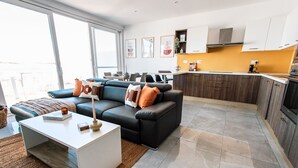 A modern living room with a black sofa, white coffee table, and a kitchen area with wooden cabinets and a countertop. - Pretty Beachfront Penthouse in Mellieha (Il-Mellieha)