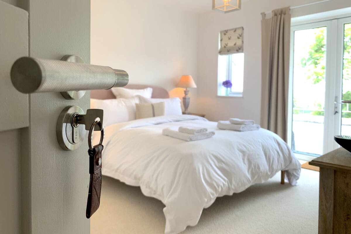 Quarto de hotel com cama, mesa de cabeceira, luminária, janela com cortinas e vista para o verde lá fora.