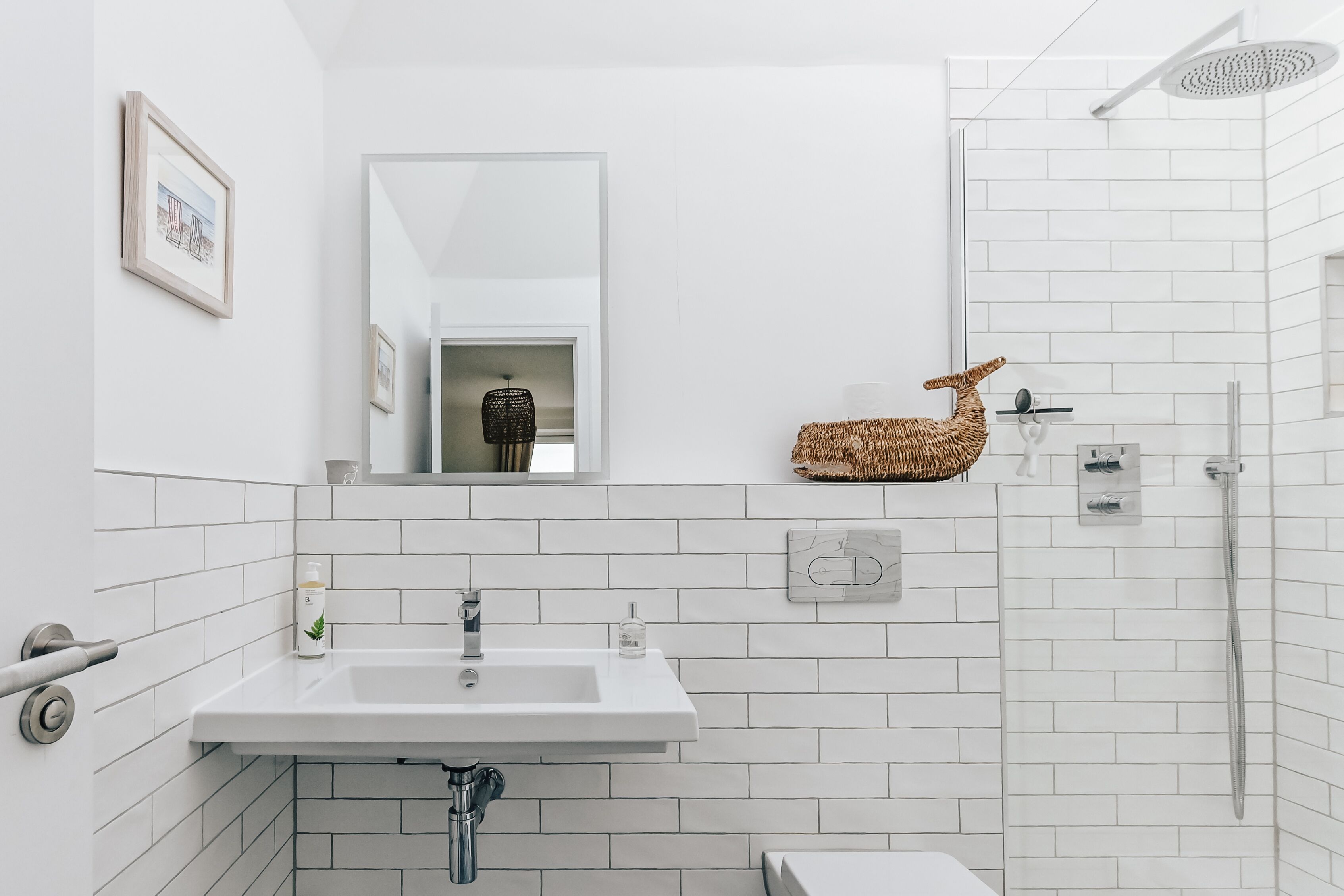 A modern bathroom with white tiles, a sink, a mirror, a shower, and a decorative basket.