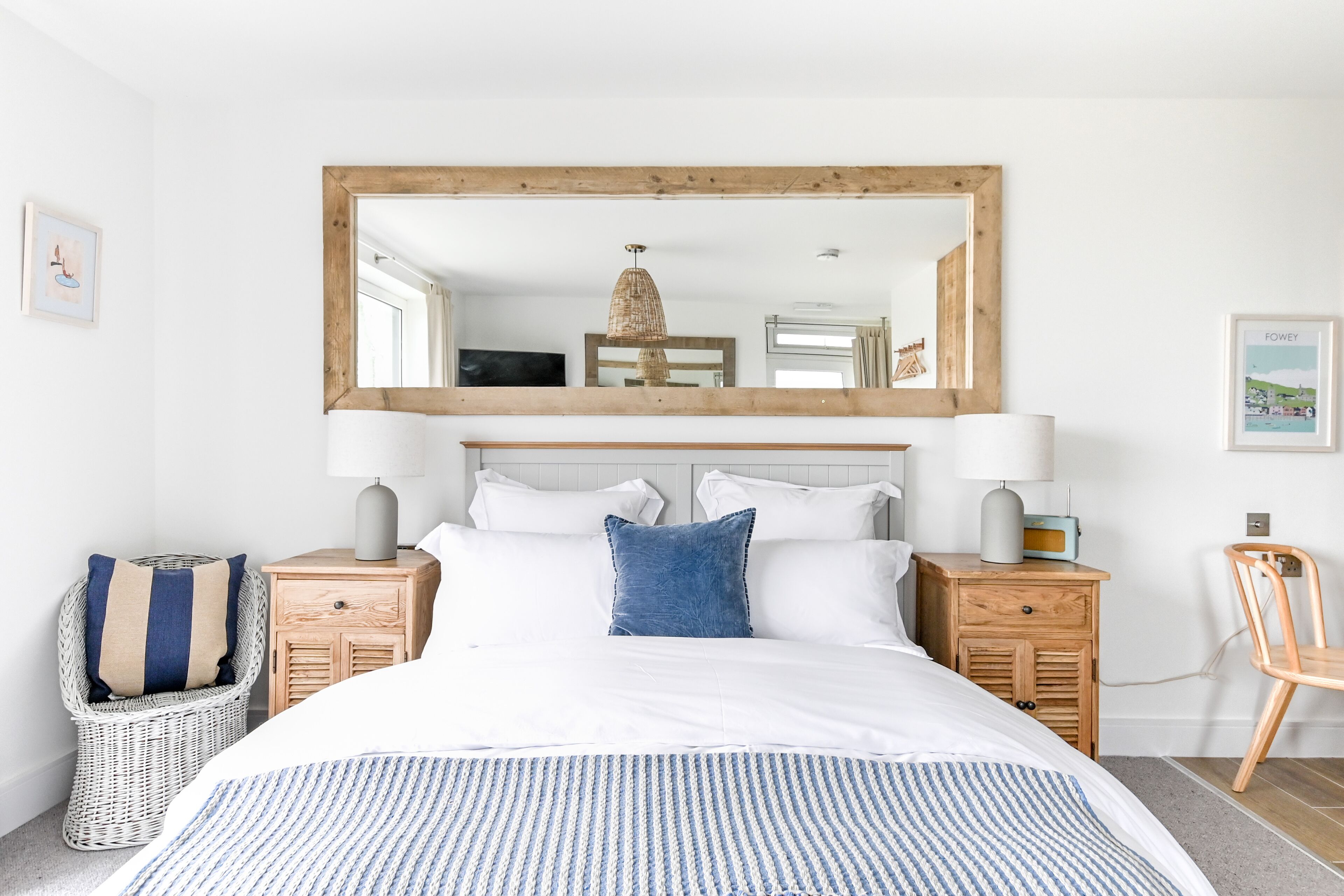 A bedroom with a large bed, two bedside tables, a wicker chair, a wooden chair, and a large mirror on the wall.