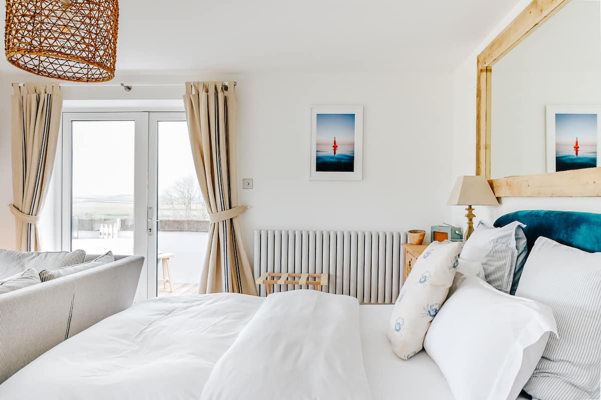 Quarto de hotel com cama, cabeceira de madeira, radiador, luminária e janela com cortinas.
