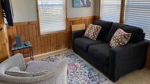 Ein gemütliches Wohnzimmer mit einer dunklen Couch, gemusterten Kissen, einem kleinen Beistelltisch und einem farbenfrohen Teppich.