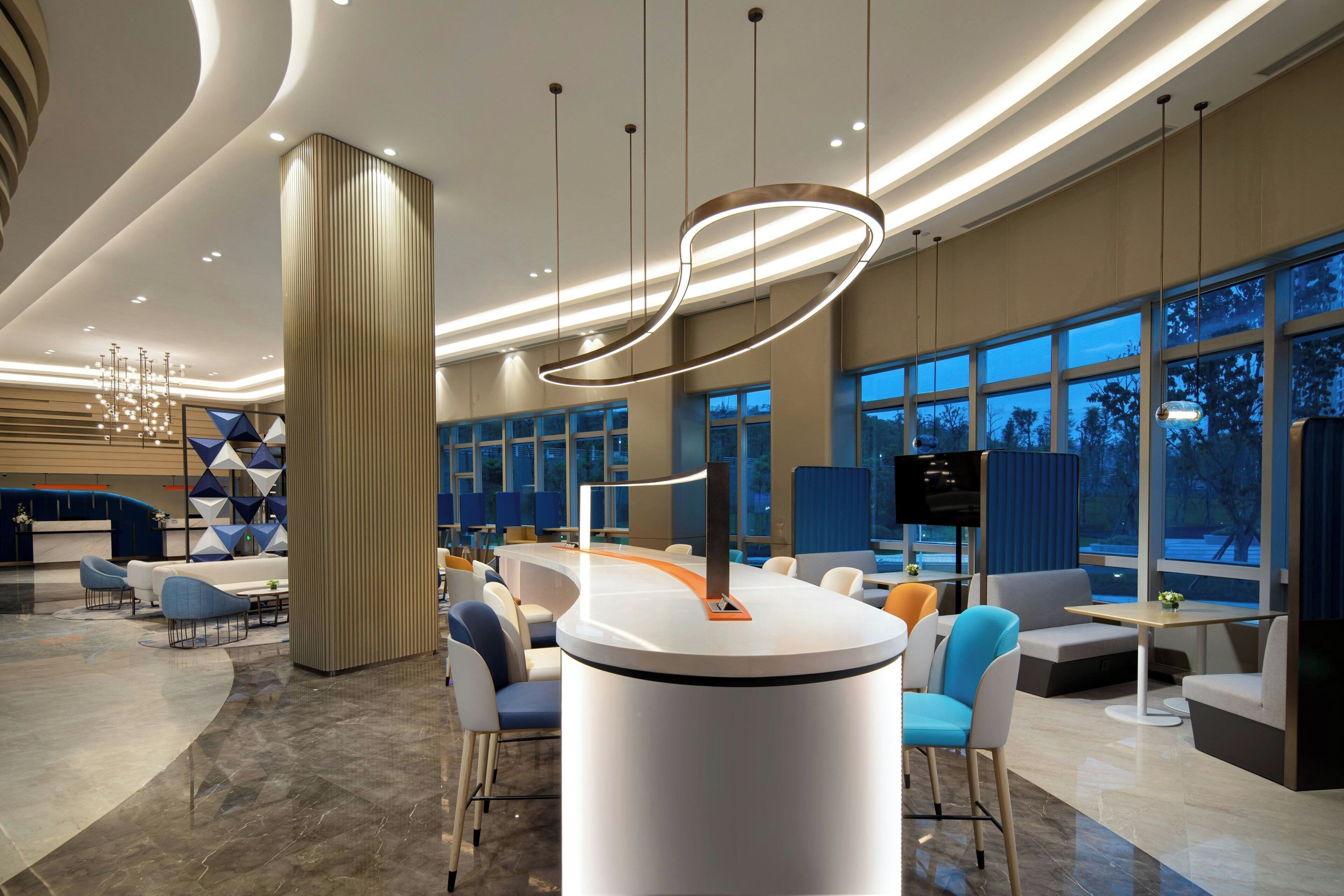A modern hotel lobby with a central reception desk, seating areas, and large windows.