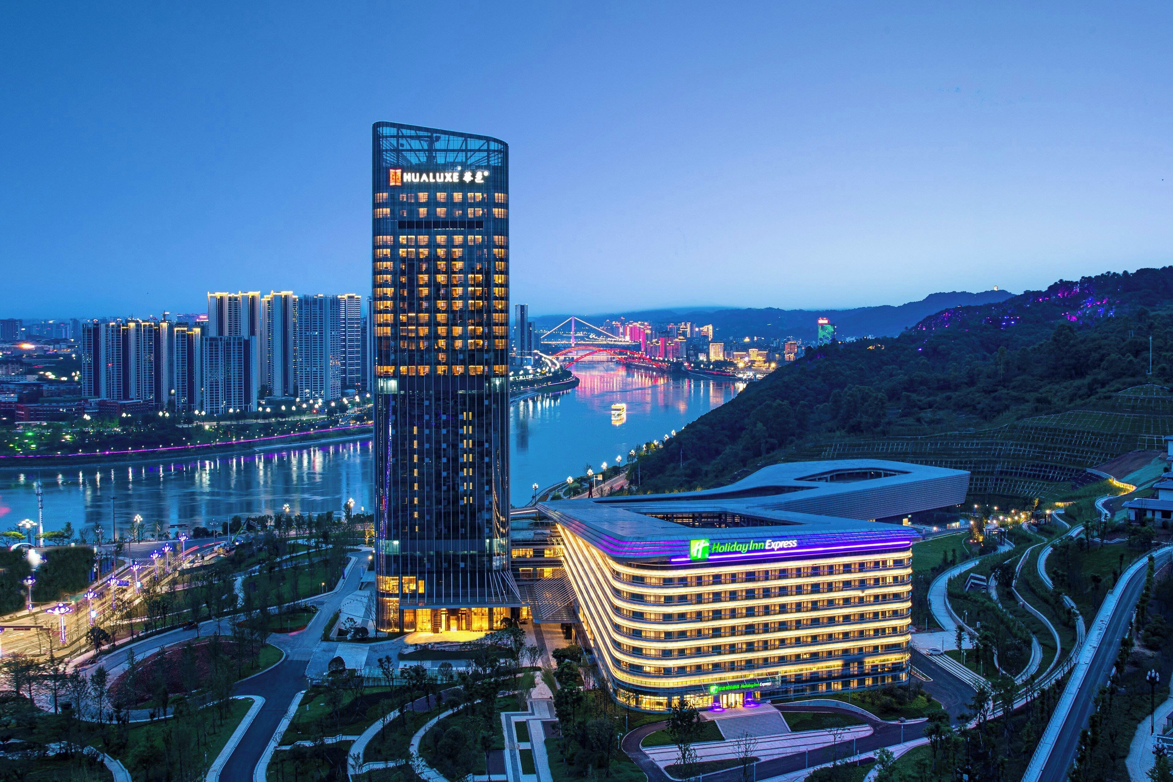 A modern hotel with a curved facade, located near a river with a bridge in the background.