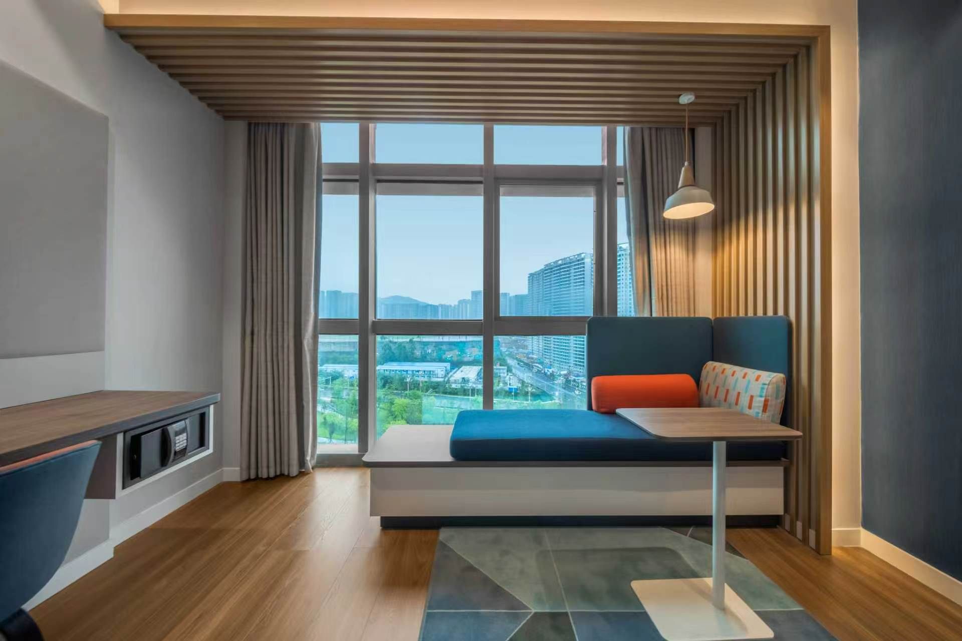 Une chambre moderne dotée d’une grande fenêtre, d’une banquette intégrée avec des coussins, d’un bureau et d’un luminaire suspendu.