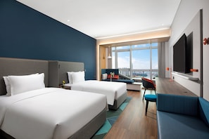 Two large beds with grey headboards and white bedding, a sofa, and a flat-screen TV in a modern hotel room with a city view.