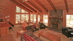 A cozy cabin interior with a stone fireplace, wooden furniture, and large windows offering a view of the forest. - Cozy 3 bed, 2 bath, Pet Friendly home. HOT TUB! (Black Butte Ranch)