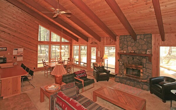 A cozy cabin interior with a stone fireplace, wooden furniture, and large windows offering a view of the forest.