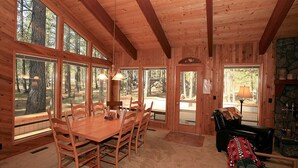 A wooden cabin interior with a large dining table, multiple chairs, and large windows offering views of the surrounding forest. - Cozy 3 bed, 2 bath, Pet Friendly home. HOT TUB! (Black Butte Ranch)