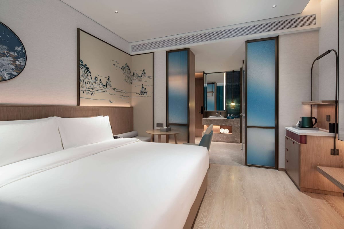 A modern hotel room with a large bed, a small round table, a mirror, and a kitchenette area.