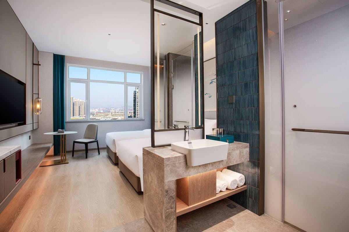 A modern hotel room with a large bed, a sitting area, a bathroom with a large mirror, and a view of the city.