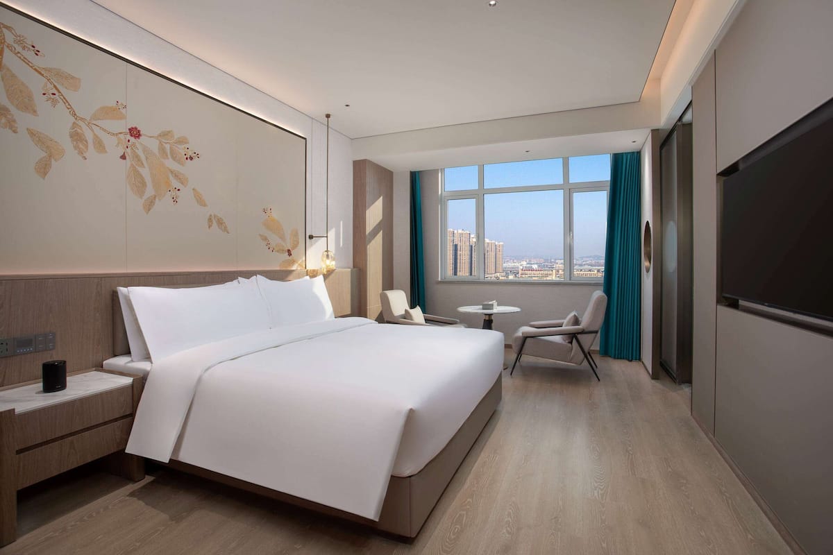 A modern hotel room with a large bed, bedside tables, a desk, and a view of the city through large windows.