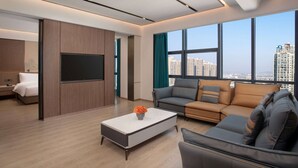 A modern living room with a flat-screen TV, a sofa, a coffee table, and a large window offering a city view. - Ramada by Wyndham Huangshi Huangshigang (Huangshi)