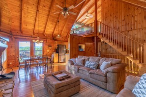 A spacious log cabin interior with a dining area, a sofa, and a staircase. - Whispering Ridge Retreat - 7 mins. to downtown Boone, hot tub, great views, pool table! (Boone)