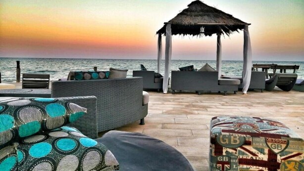 Un salon de plage doté d’un gazebo à toit de chaume, de chaises longues et de coussins.