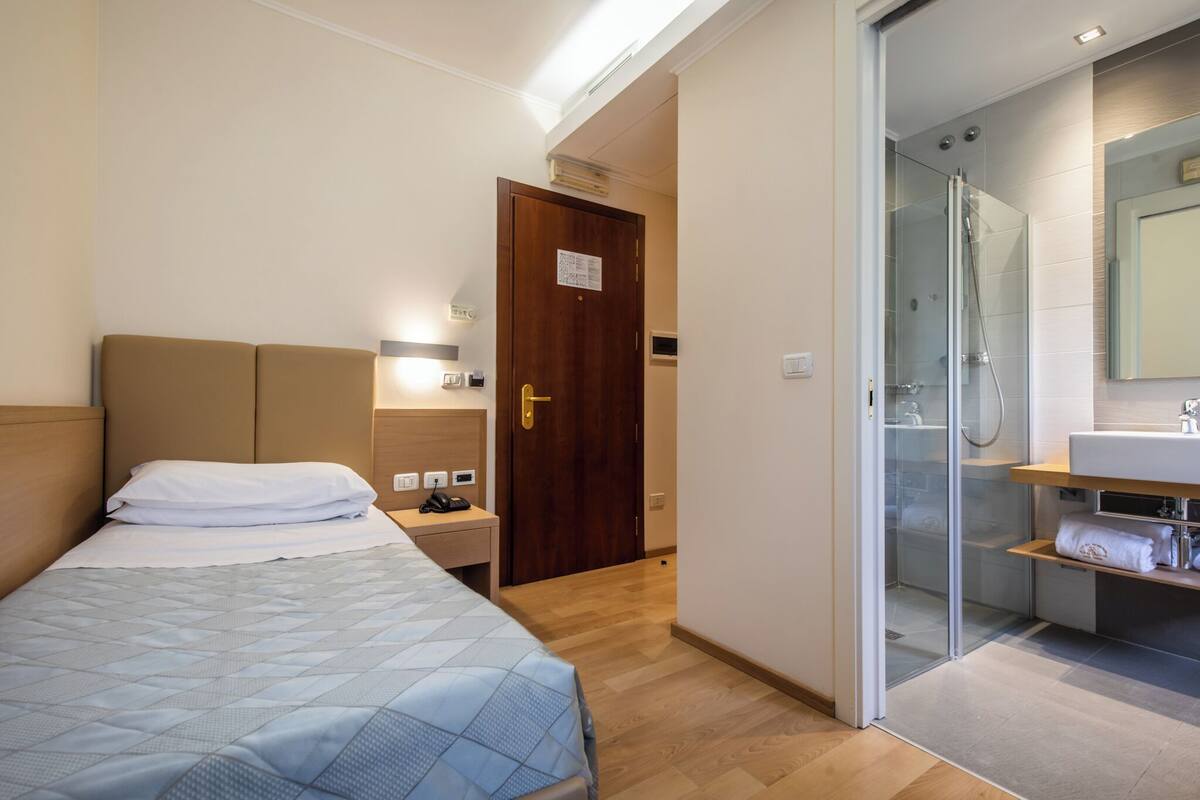 Habitación de hotel con cama, mesita de noche, lámpara y zona de ducha con mampara de vidrio.