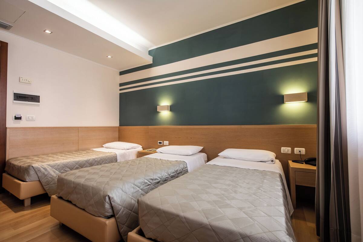 Habitación de hotel con tres camas, cabecera de madera, luces fijadas a la pared y mesita de noche con teléfono.