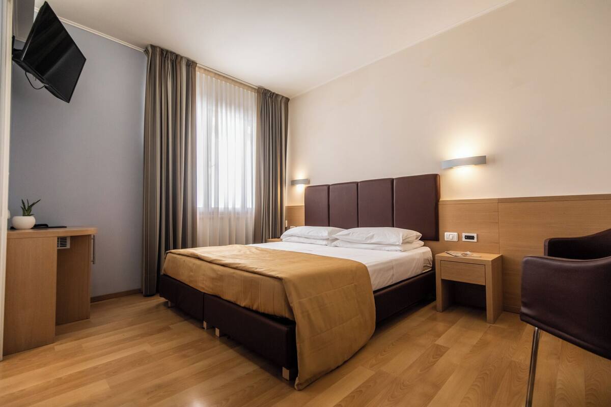 Una habitaci ón de hotel con cama, mesitas de noche, escritorio, silla, televisor montado en la pared y ventana con cortinas.