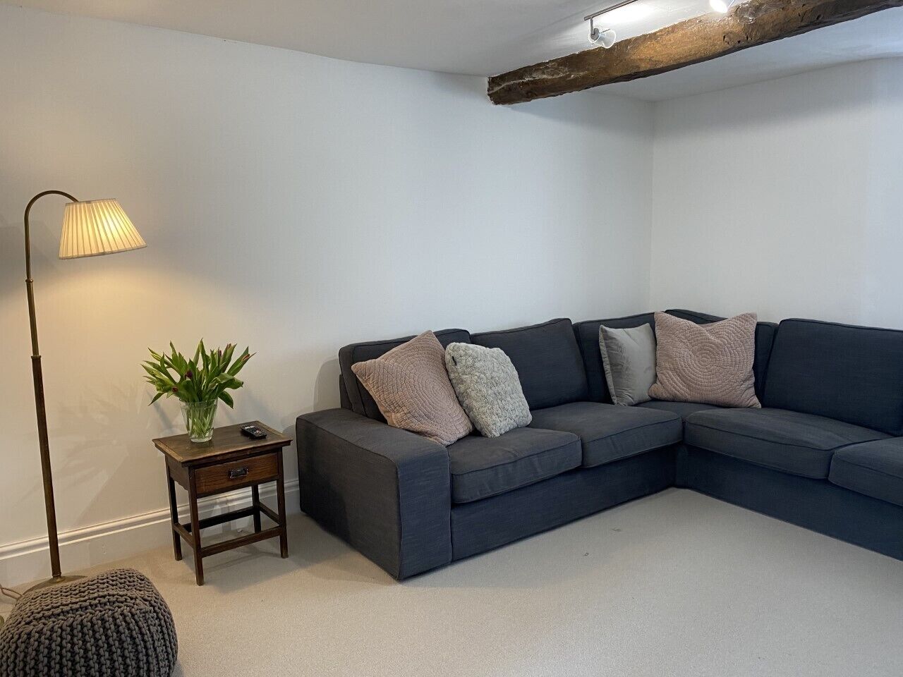 Een moderne woonkamer met een donkergrijze hoekbank, een klein houten bijzettafeltje met een lamp en een potplant.