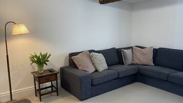 Een moderne woonkamer met een donkergrijze hoekbank, een klein houten bijzettafeltje met een lamp en een potplant.