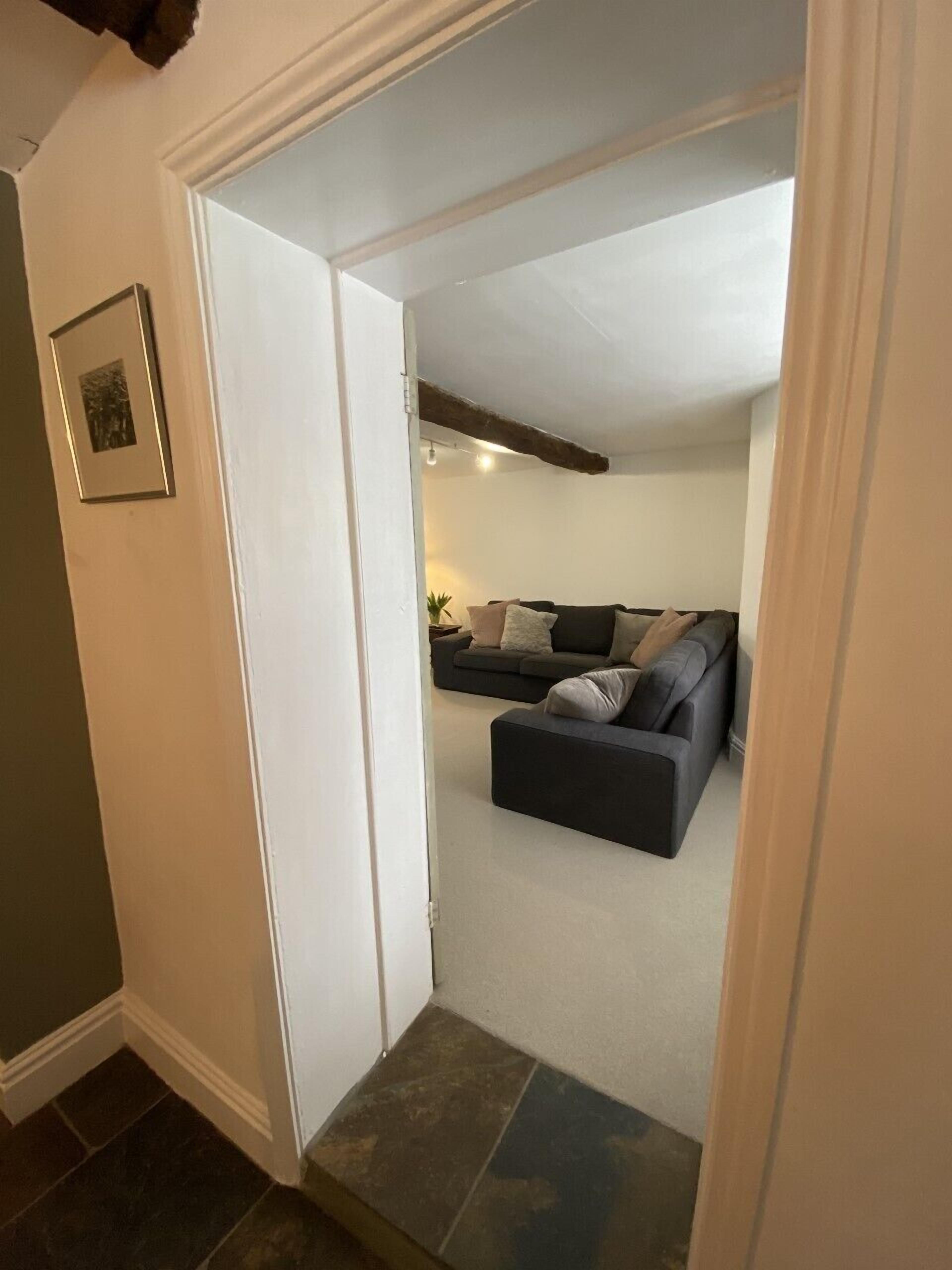 A narrow hallway leading to a living area with a dark-colored sofa set and a small table with a lamp.