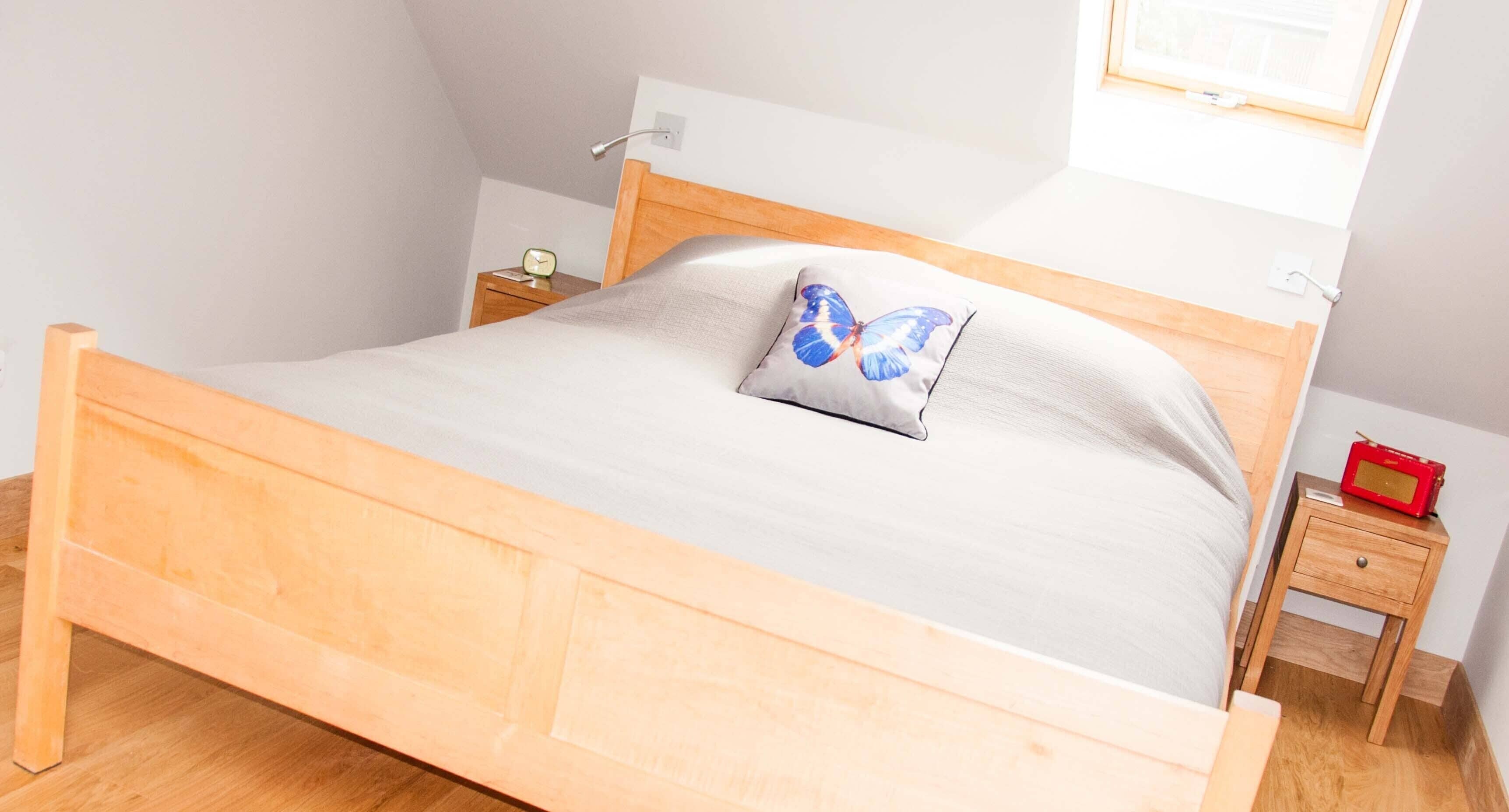 Una cama de madera con un colchón gris y un almohadón con un diseño de mariposa azul. Hay una mesita de noche de madera con una radio roja y un reloj pequeño.