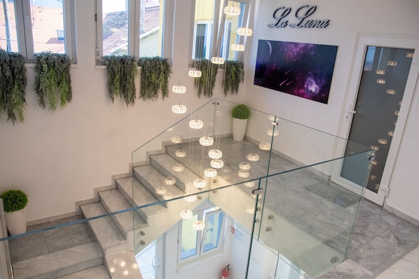 A modern hotel interior with a glass staircase, hanging plants, and a large wall-mounted TV displaying a space-themed image.