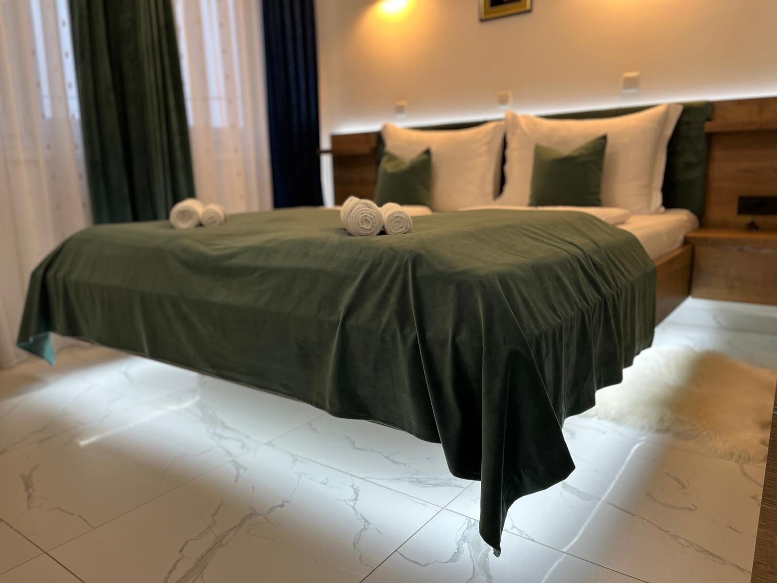 Un lit bien fait, recouvert d’une couvre-lit verte et agrémenté de deux serviettes enroulées, avec une tête de lit en bois et un sol en marbre blanc.