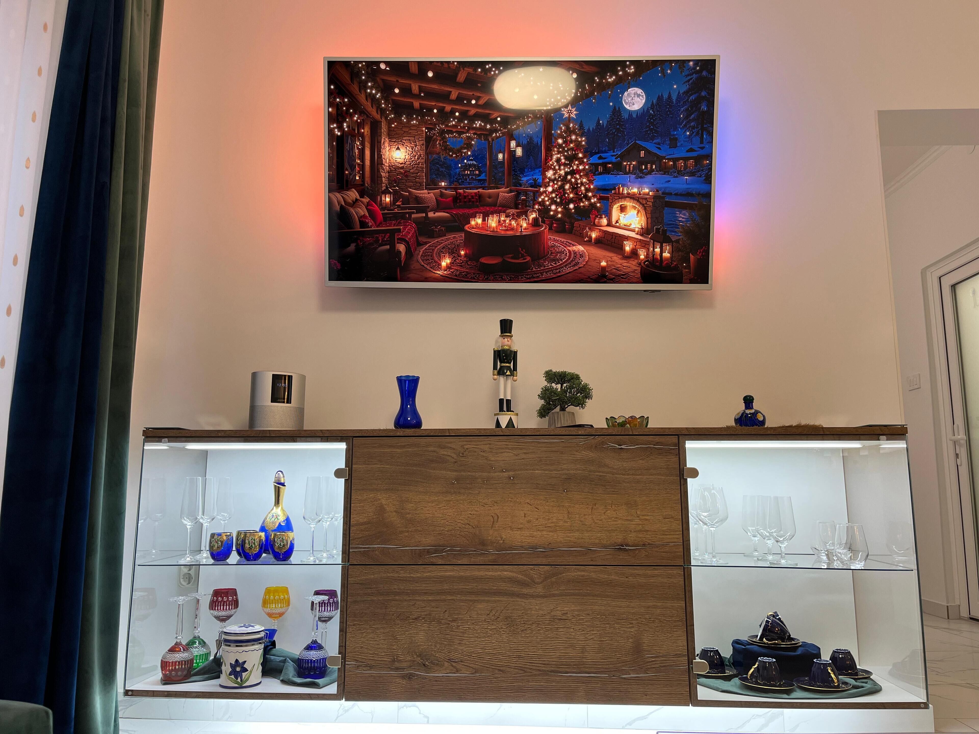A living room with a TV mounted on the wall displaying a winter scene. Below the TV, there is a wooden cabinet with glass shelves displaying various glassware.