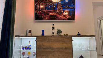 A living room with a TV mounted on the wall displaying a winter scene. Below the TV, there is a wooden cabinet with glass shelves displaying various glassware.
