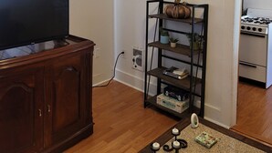 A living room with a TV on a wooden cabinet, a black metal shelf with plants, a white stove, and a painting on the wall. - Stylish Retreat w/ Queen Bed • Fast WiFi • Free Parking (Colorado Springs)