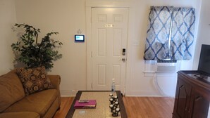 A living room with a brown sofa, a coffee table with books and a bottle, a potted plant, a door with a no smoking sign, and a patterned curtain. - Stylish Retreat w/ Queen Bed • Fast WiFi • Free Parking (Colorado Springs)