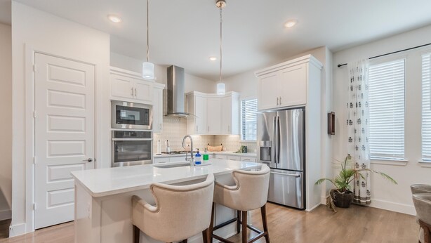 A modern kitchen with white cabinetry, stainless steel appliances, a central island with bar stools, and a large window with blinds. - Contemporary Home with Office, Centrally Located (Plano)