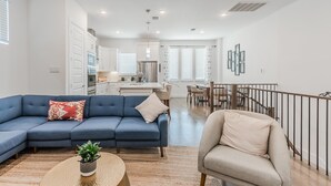 A modern living room with a blue sofa, a beige armchair, and a round coffee table with a plant. A kitchen and dining area are visible in the background. - Contemporary Home with Office, Centrally Located (Plano)
