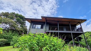 Een modern huis met twee verdiepingen en een balkon, omgeven door weelderige begroeiing.