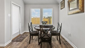 Un comedor con una mesa redonda y cuatro sillas, dos ventanas con persianas y una puerta.