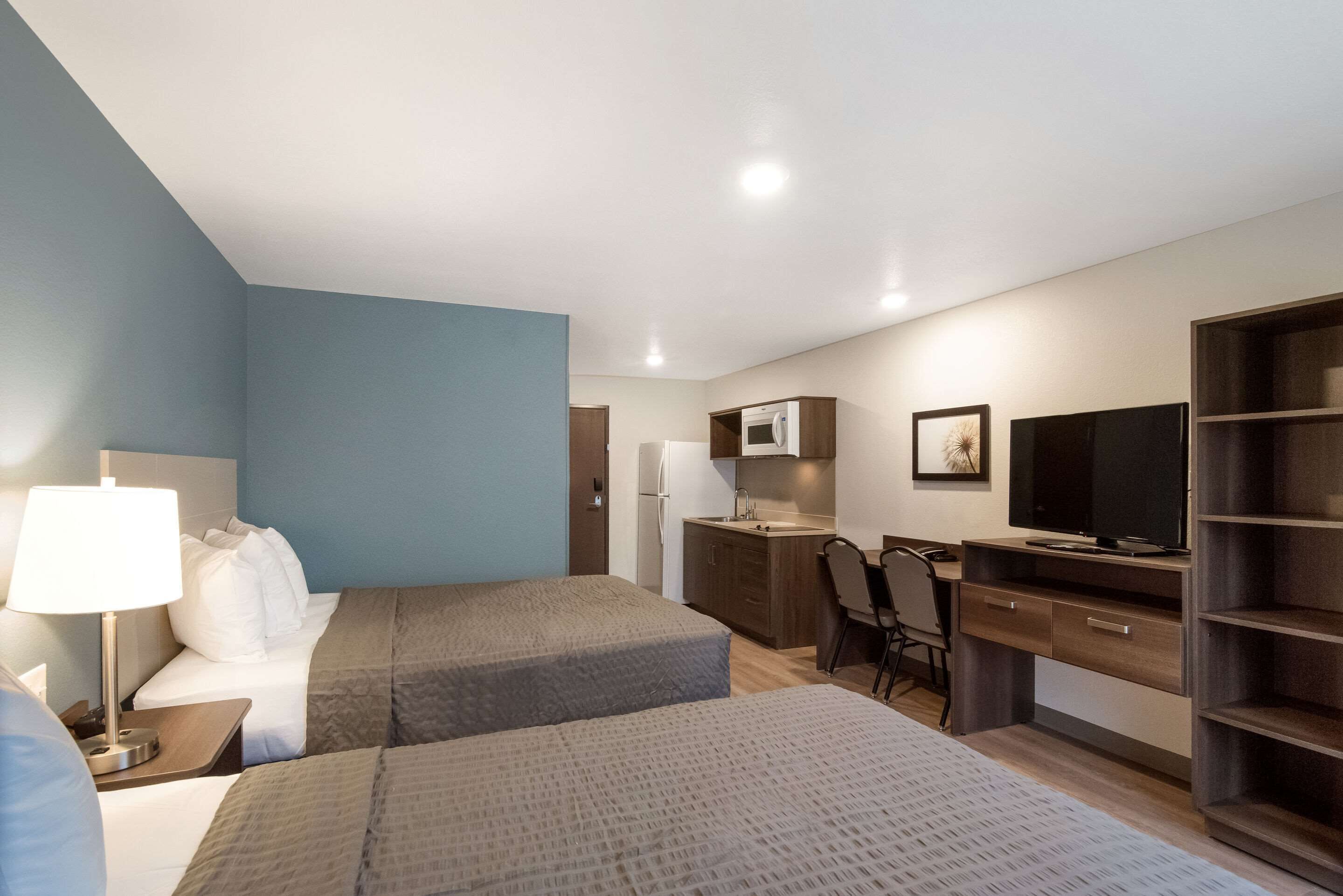 A hotel room with two beds, a desk, a chair, a TV, a wardrobe, and a kitchenette.