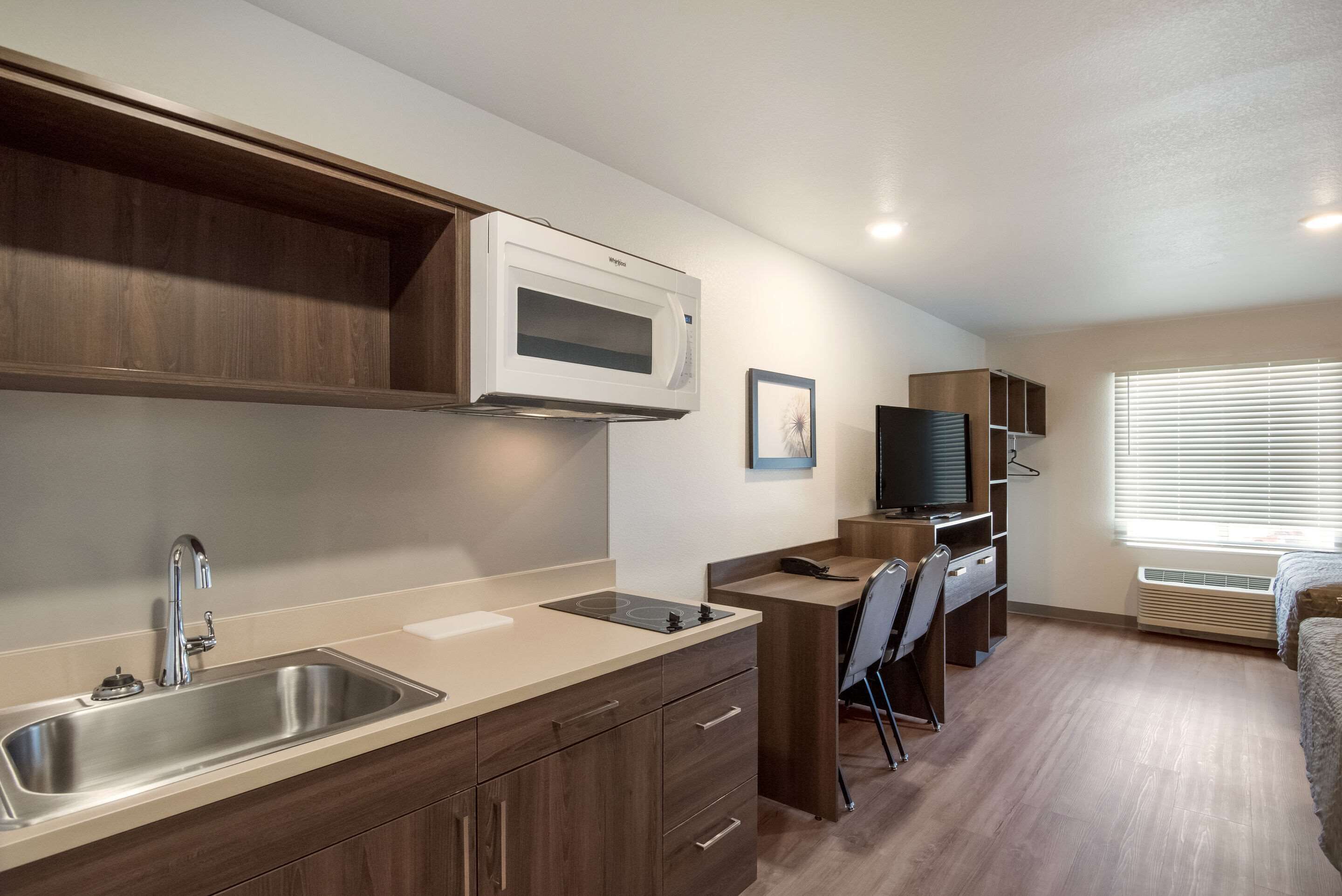 Una kitchenette compacta con microondas, pileta y estufa, un escritorio pequeño con una silla, y un televisor.