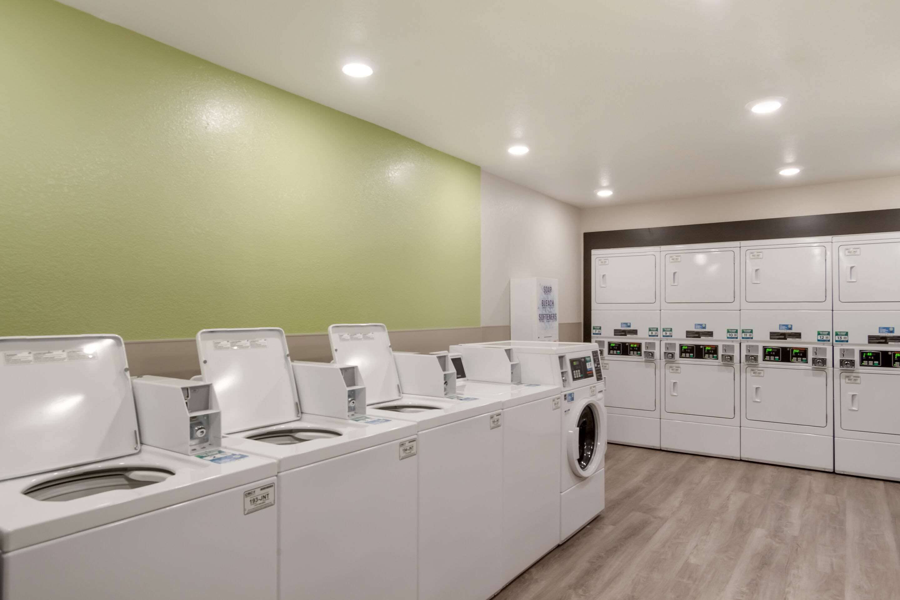 A laundry facility with multiple washing machines and dryers, a green wall, and a wooden floor.