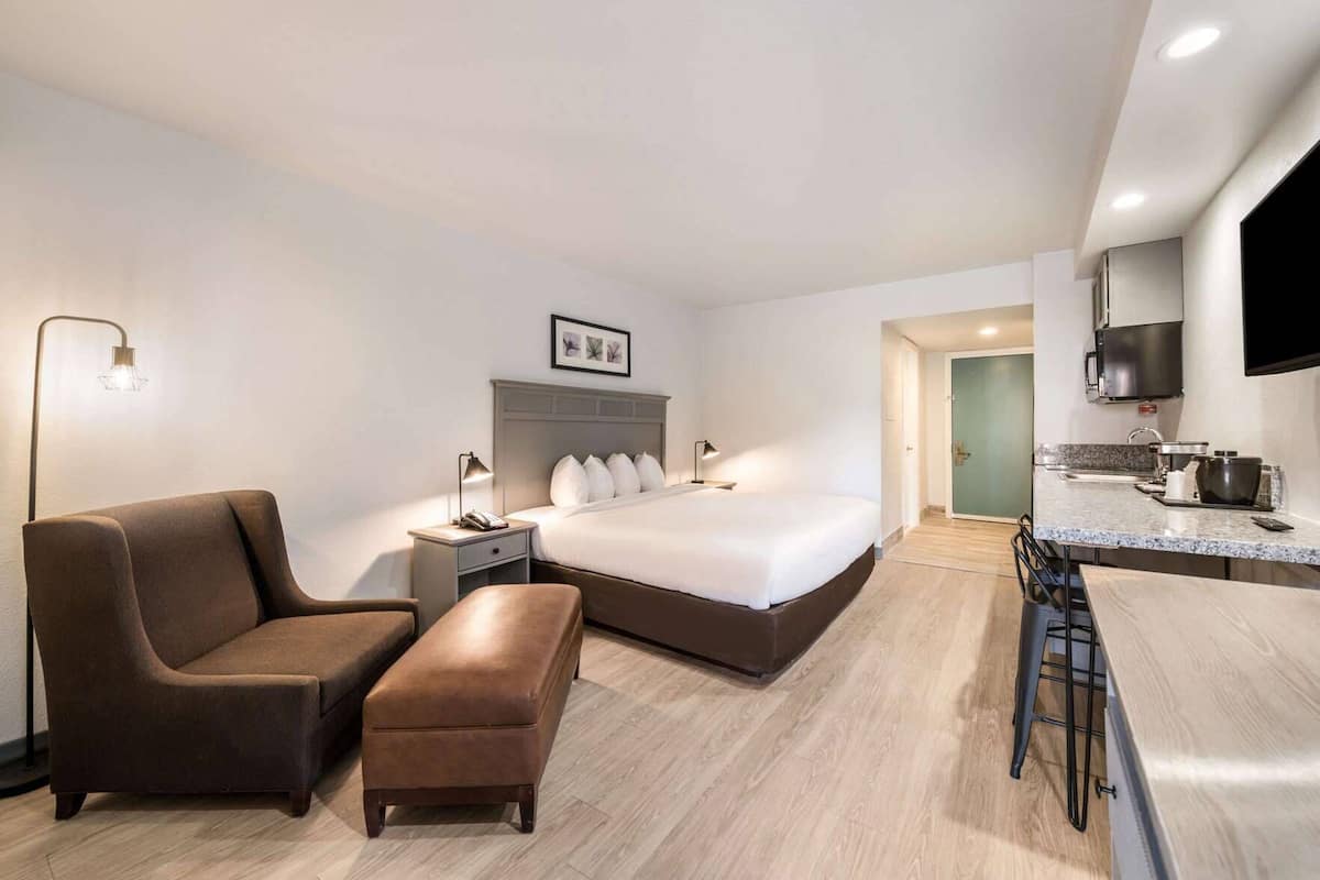 A modern hotel room with a bed, a desk, a chair, and a kitchenette.