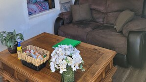 Un salon chaleureux avec un canapé marron, une table basse en bois agrémentée d’un panier et de fleurs, une fenêtre avec des rideaux à motifs et un tableau accroché au mur.