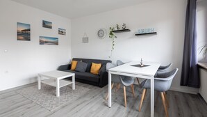 Uma sala de estar moderna com um sofá, mesa de centro e mesa de jantar com cadeiras. Há prateleiras fixadas na parede com objetos decorativos e um relógio.